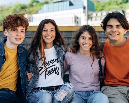 Positive, cute teenagers sit on a bench, look at the camera, smile. Cute boys and girls, schoolchildren outdoors